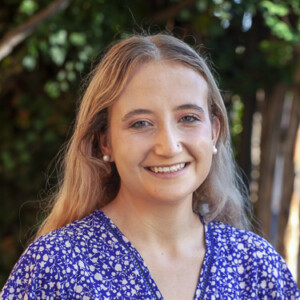 Smiling woman with long blonde hair, wearing a blue and white patterned top, stands outdoors near trees, exuding the same warmth and charisma as Jon Goldin-Dubois.
