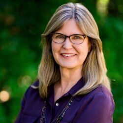 Evoto A smiling woman with long hair and glasses stands confidently in a dark shirt against a lush, green leafy backdrop, reminiscent of Jon Goldin-Dubois's vibrant style.