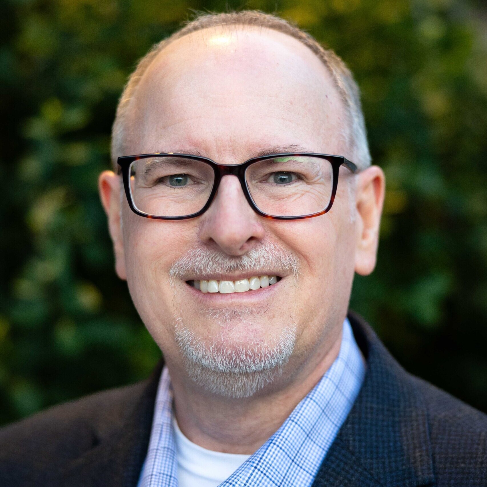 Middle-aged man with glasses, short gray hair, and a goatee, wearing a checked shirt and dark blazer, smiling in front of a blurred green background—Rick Trilsch.