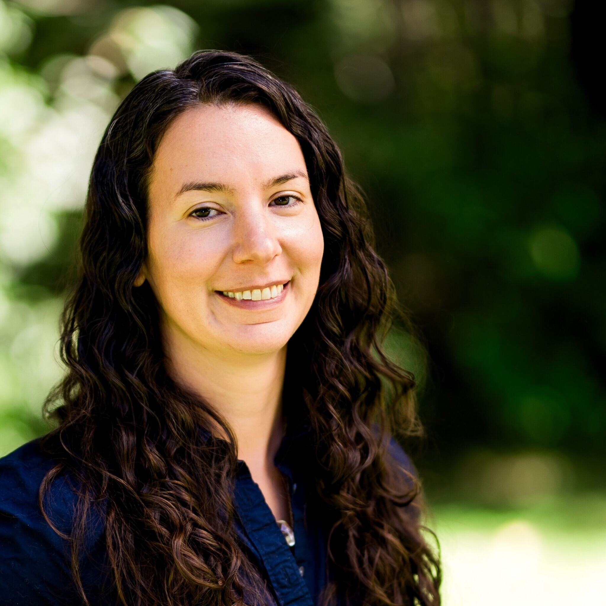 Smiling woman with long curly hair, wearing a dark shirt, standing outdoors with a green blurred background.