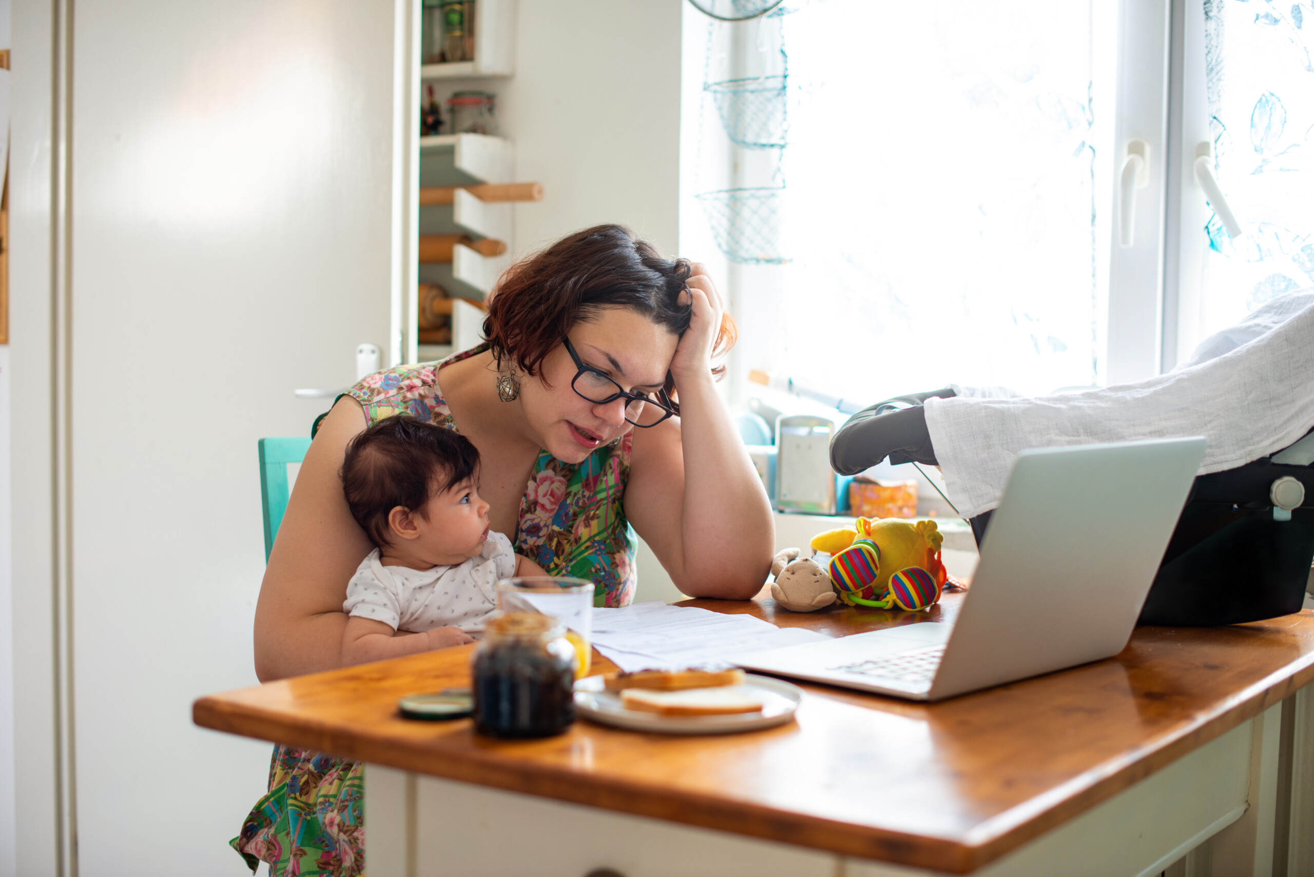 A woman sits at a table with a baby on her lap, looking at documents near a laptop. The table has toys, a jar, and a plate with food. She appears focused and thoughtful.