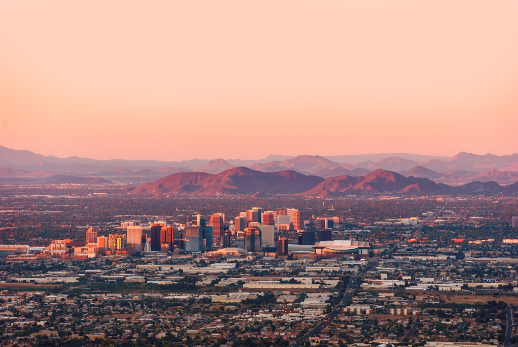 Aerial view of a cityscape with tall buildings at sunset, mountains in the background, and a pastel sky overhead.