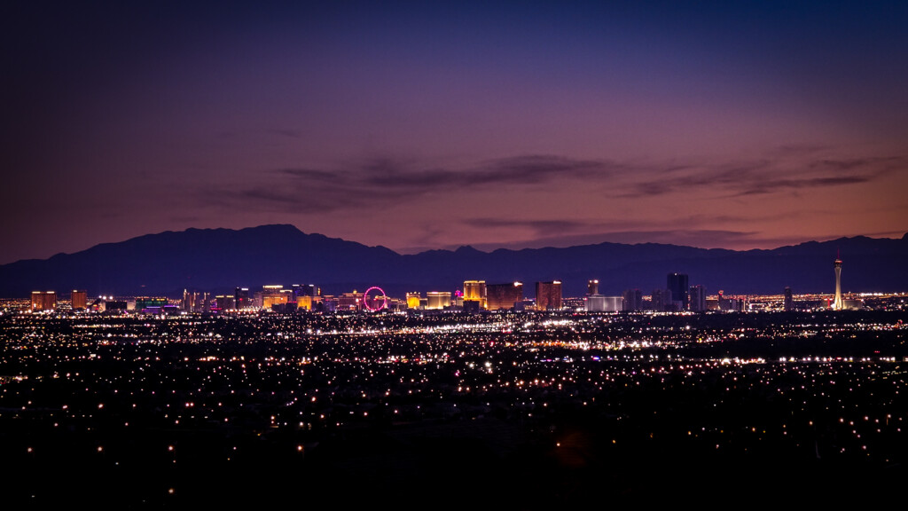 The Las Vegas Strip skyline illuminated at dusk, with the silhouette of distant mountains under a purple and orange sky.