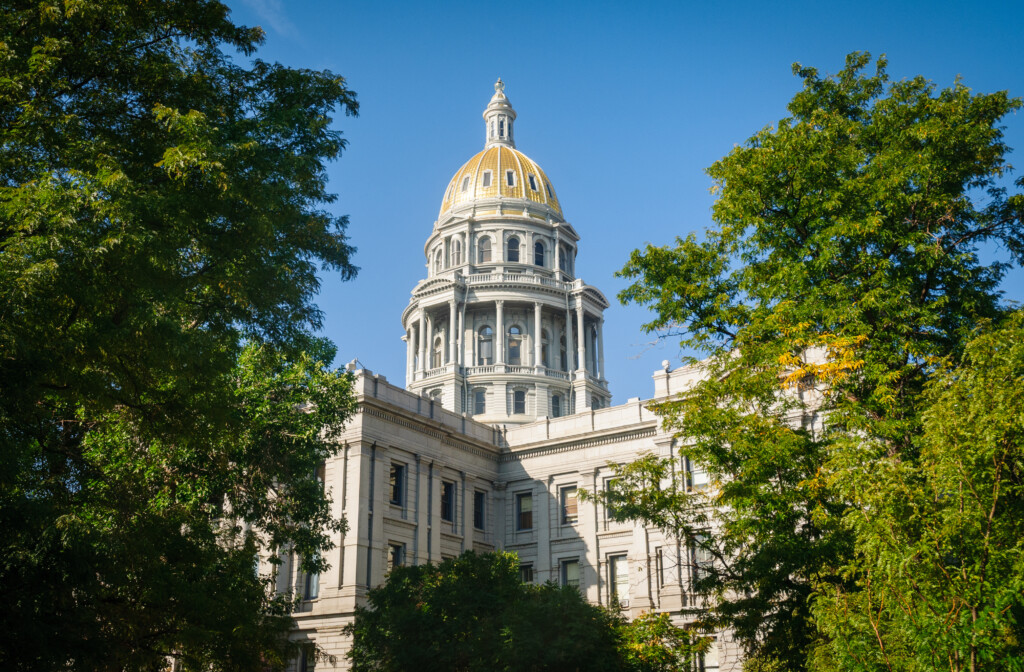 The image shows the Colorado State Capitol building with its golden dome, framed by green trees under a clear blue sky.
