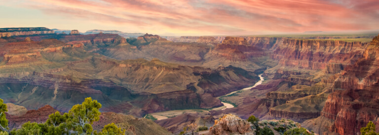 Panoramic view of the Grand Canyon at sunset with a winding river below and vibrant pink and orange sky above.