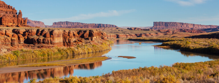 A winding river flows through a desert landscape with red rock formations under a clear blue sky. Bushes and shrubs line the riverbanks.