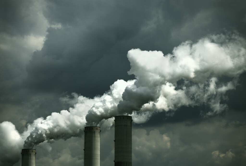 Three industrial coal smokestacks emit thick white smoke against a cloudy sky.