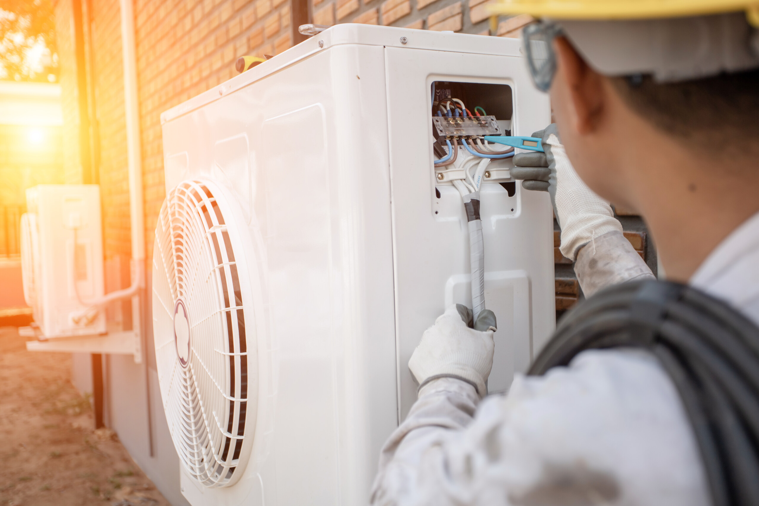 Technician in protective gear repairs an outdoor air conditioning unit against a brick wall, with sunlight in the background.