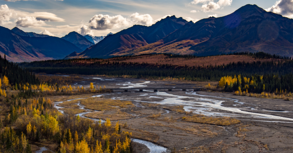 A river meanders through a wide valley with a bridge crossing, surrounded by colorful autumn trees and mountains under a partly cloudy sky.
