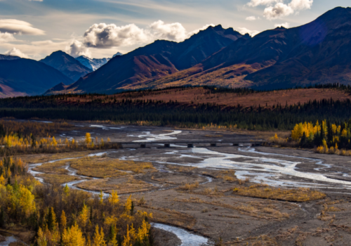 CO_Wetlands_Adobe A river meanders through a wide valley with a bridge crossing, surrounded by colorful autumn trees and mountains under a partly cloudy sky.