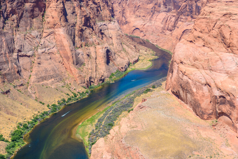 Aerial view of a winding river cutting through a canyon with reddish-brown rock formations and patches of green water and vegetation.