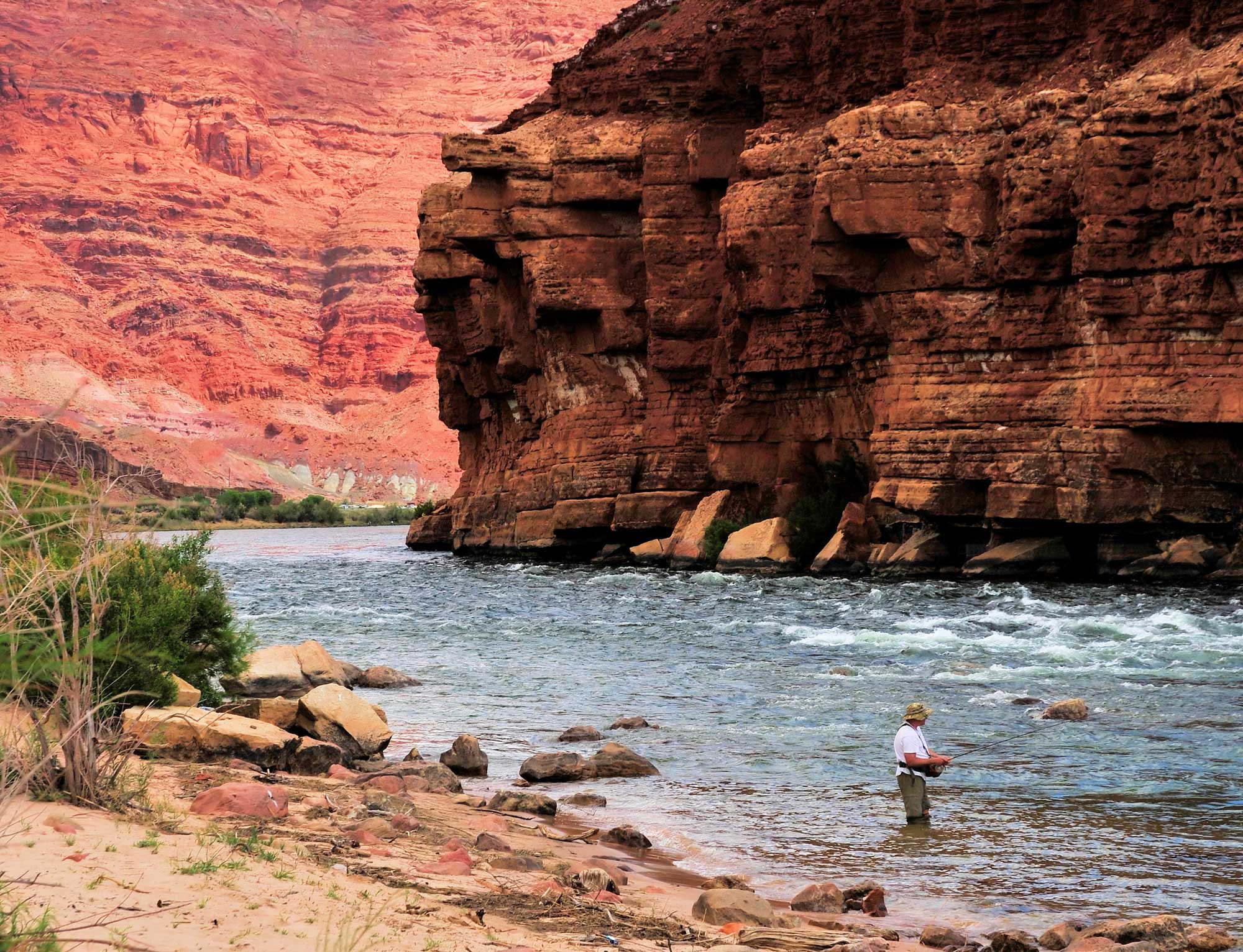 Person fishing in a river with rugged red rock cliffs in the background.