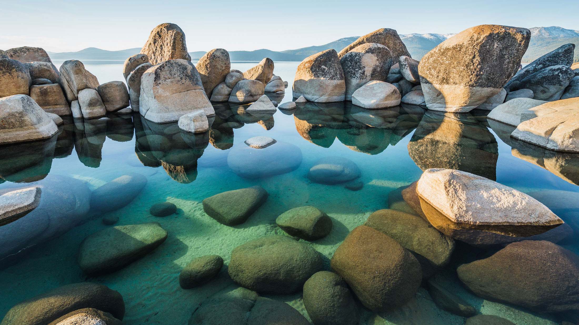 Clear water reflecting large boulders at the edge of a lake, with partially submerged rocks visible beneath the surface. Mountains can be seen in the background under a clear sky.