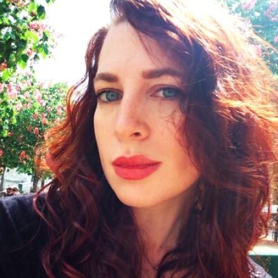 Person with long red hair and red lipstick poses outdoors with trees and flowers in the background.