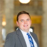 Riely Wheeler 2024 Headshot A person wearing a gray suit, white shirt, and blue floral tie stands indoors with a blurred background.