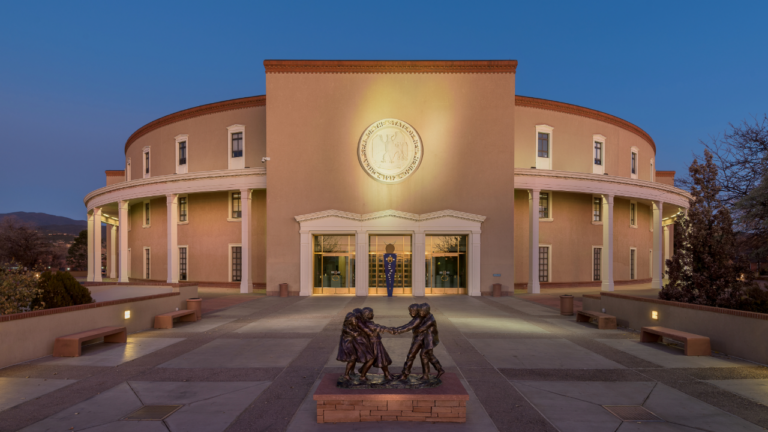 A round, brown government building with an emblem above the entrance. A bronze statue of children playing outside. Evening lighting and a clear sky.
