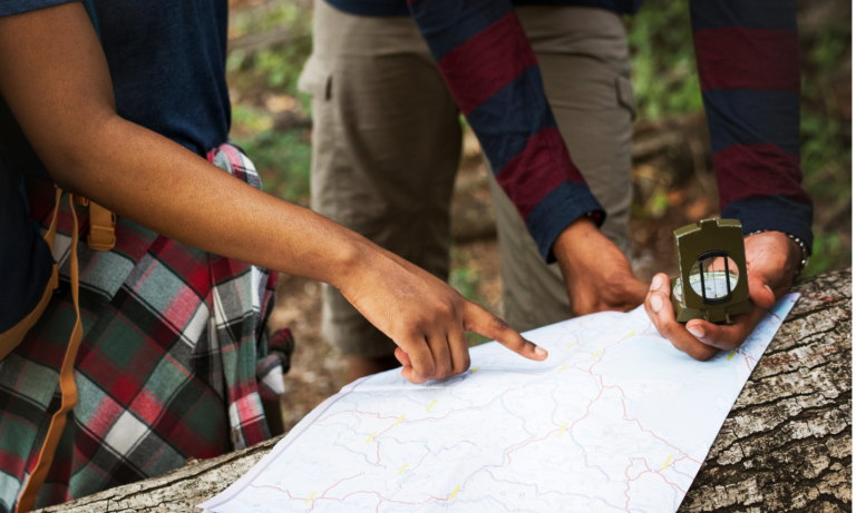 Two people in casual attire examine a map on a log in a wooded area. One points at the map, while the other holds a compass.