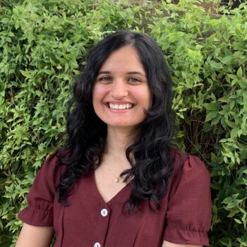 Smiling woman with long dark hair in maroon top stands in front of lush green foliage.