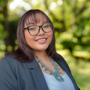 A woman with glasses and shoulder-length hair wears a blue beaded necklace and a blazer, smiling outdoors with a blurred green background.