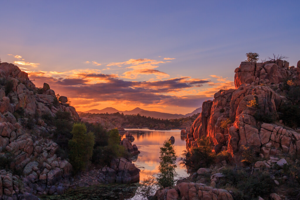 Sunset over a rocky landscape with a lake reflecting the sky. Rocky cliffs frame the scene, and distant mountains are visible under a colorful sky with orange and purple hues.