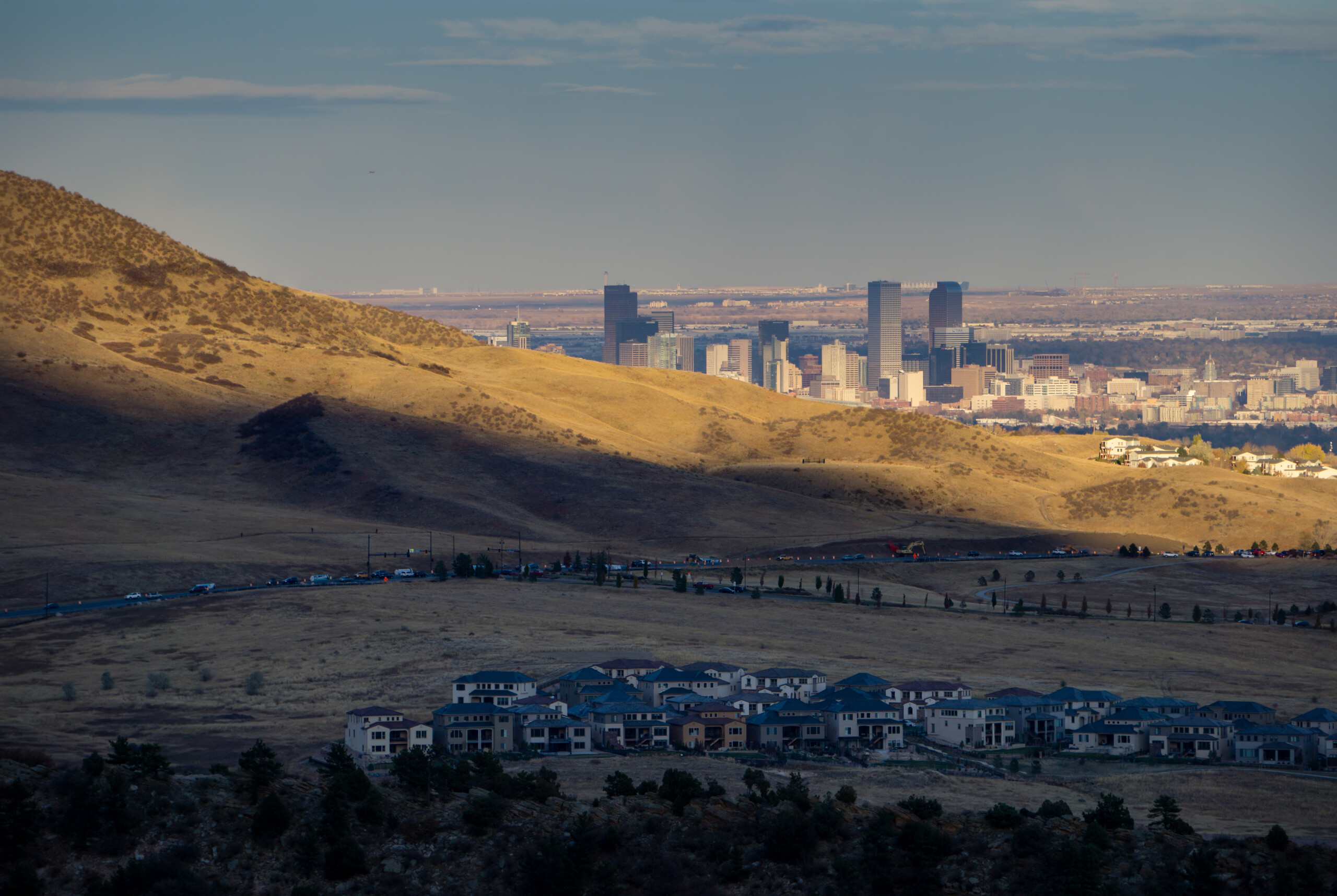 A distant view of a city skyline with tall buildings at sunset, surrounded by a hilly landscape with residential houses in the foreground.