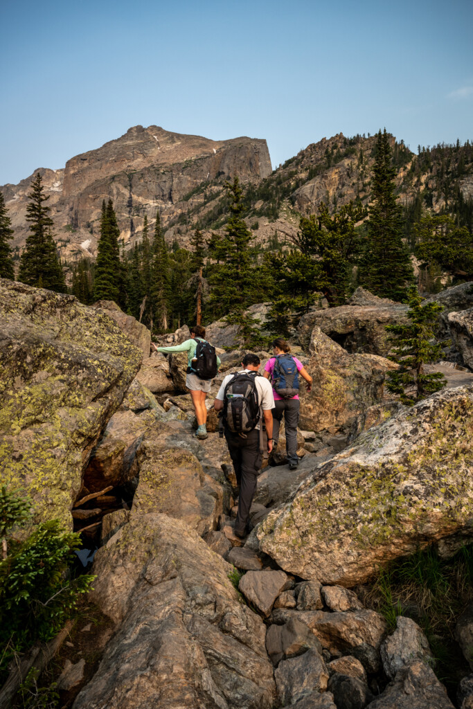 Hikers Climb Over Boulders to Haiyaha Lake Three people wearing backpacks hike on a rocky trail through a forested mountain landscape under a clear sky.