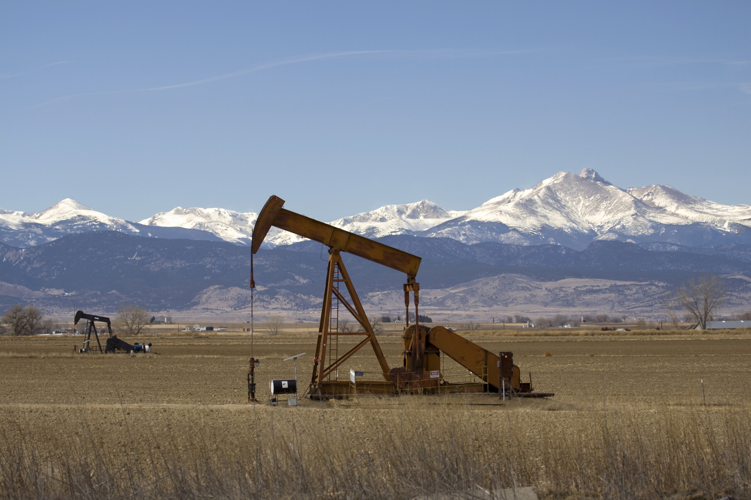 Pair of oil pumpers and Rocky Mountains, Colorado Oil pumps operate in an open field with snow-capped mountains in the background under a clear blue sky.