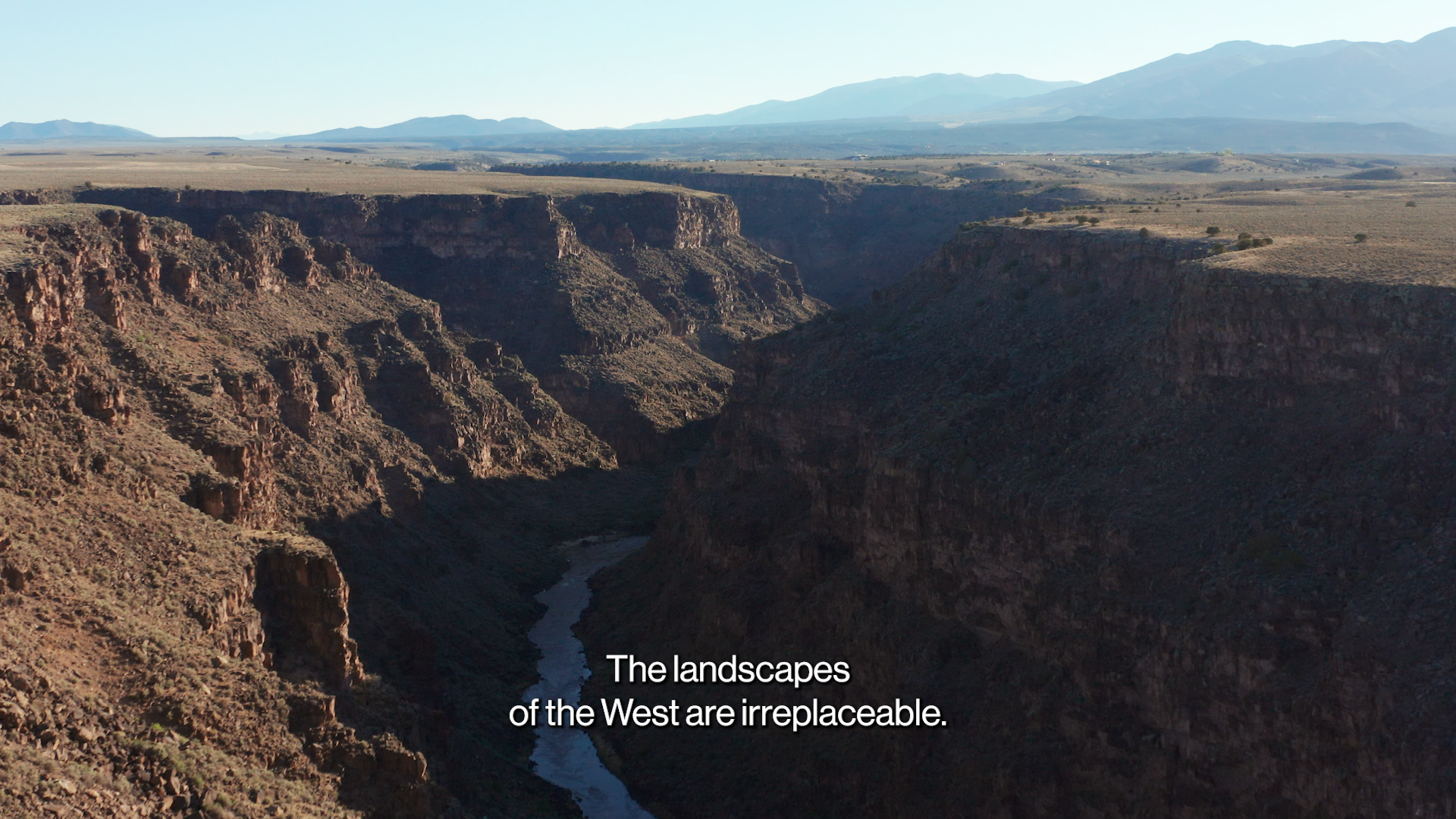 A vast canyon with a river at the bottom, surrounded by rugged cliffs and distant mountains under a clear sky. Subtitles say, "The landscapes of the West are irreplaceable.