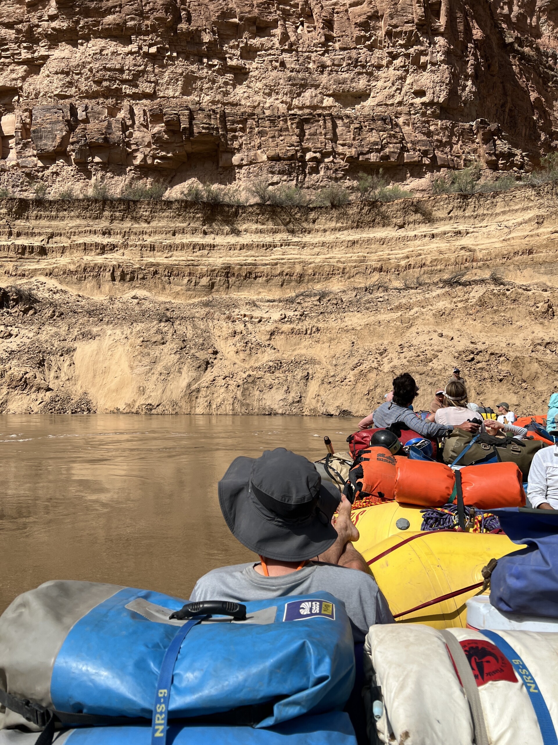 People in a raft with gear navigate a river surrounded by rocky canyon walls.