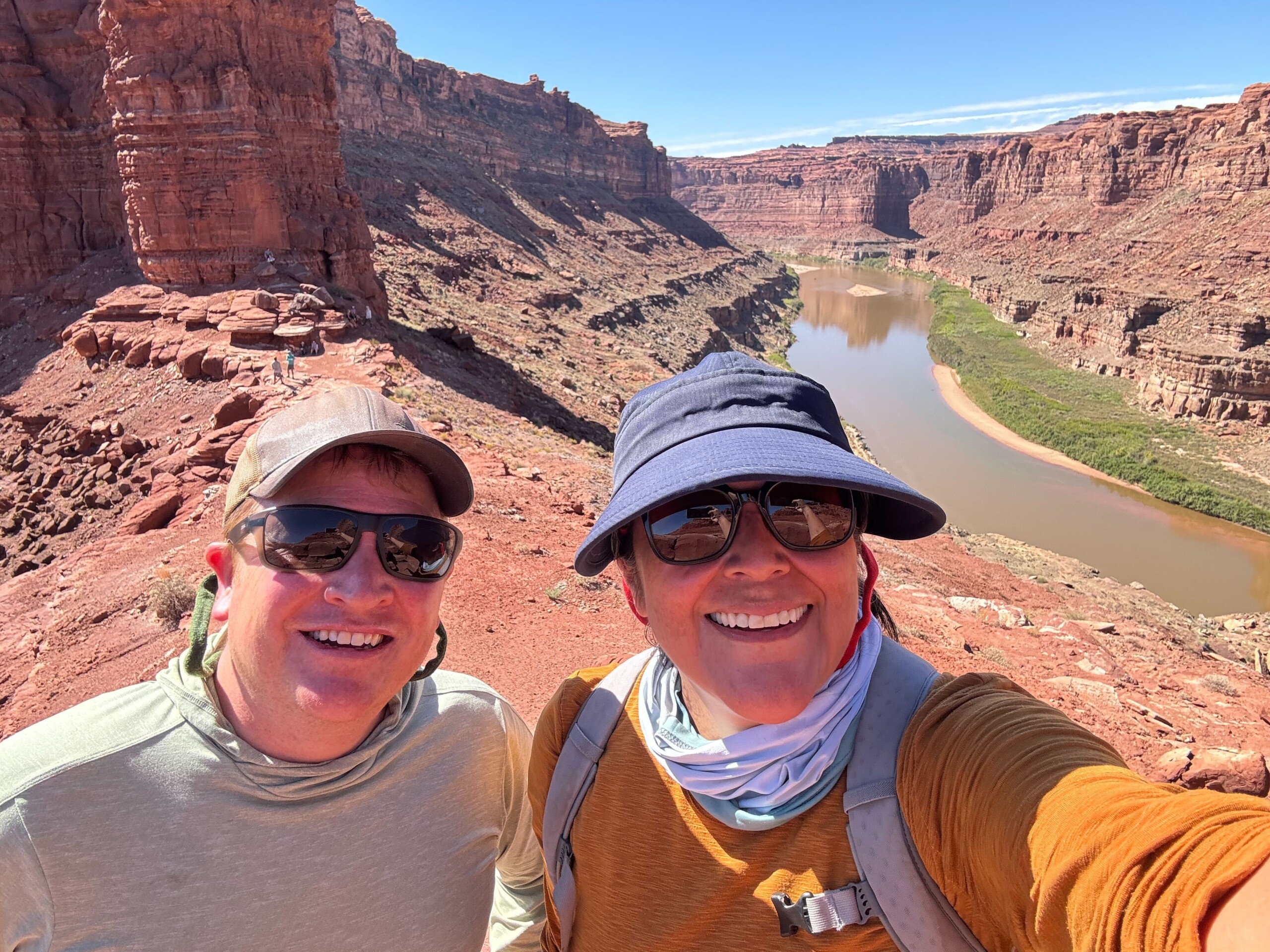 Two people wearing hats smile for a selfie on a rocky canyon cliff overlooking a river under a clear blue sky.