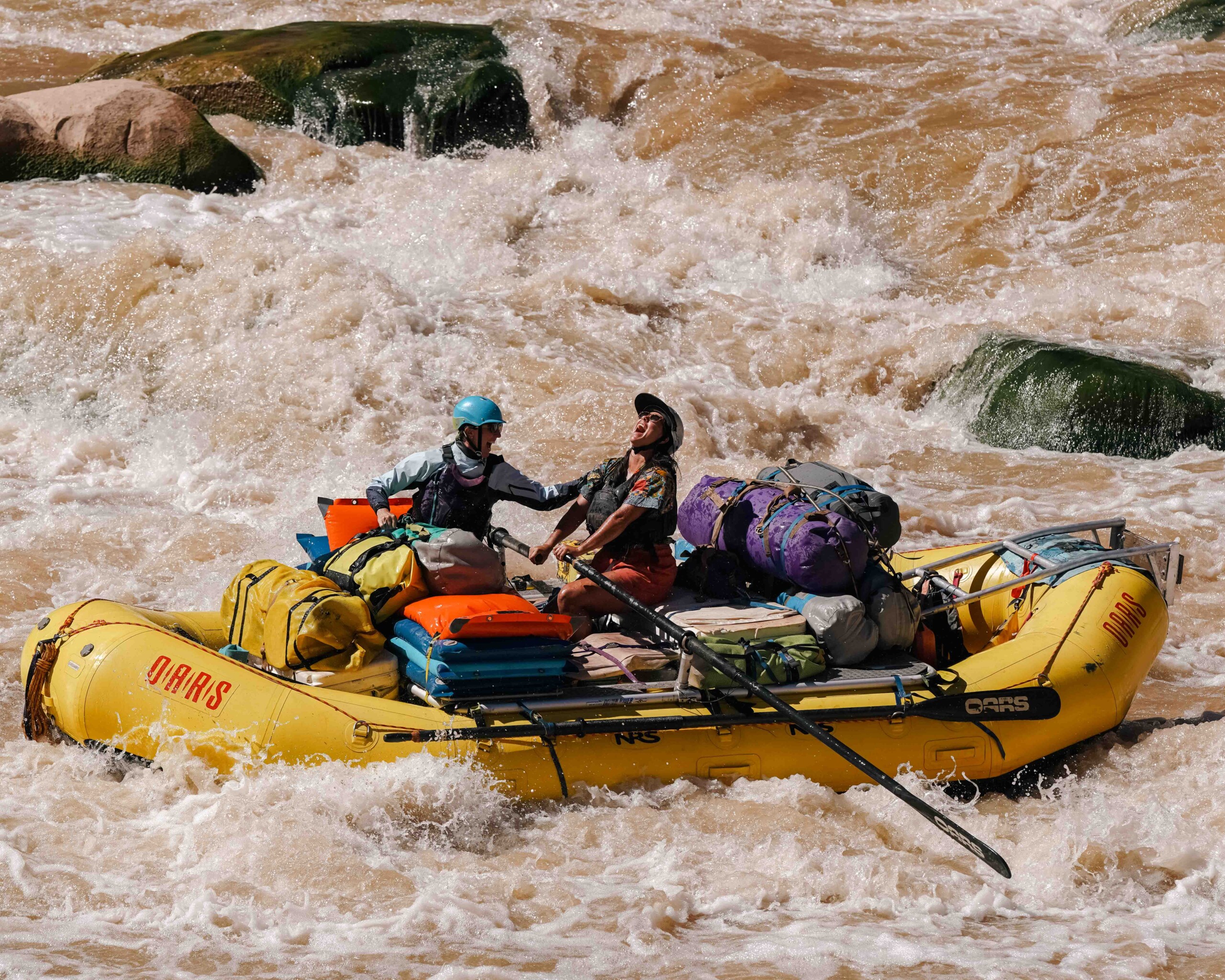 Two people navigating a yellow raft through rapids, surrounded by gear and rocks.