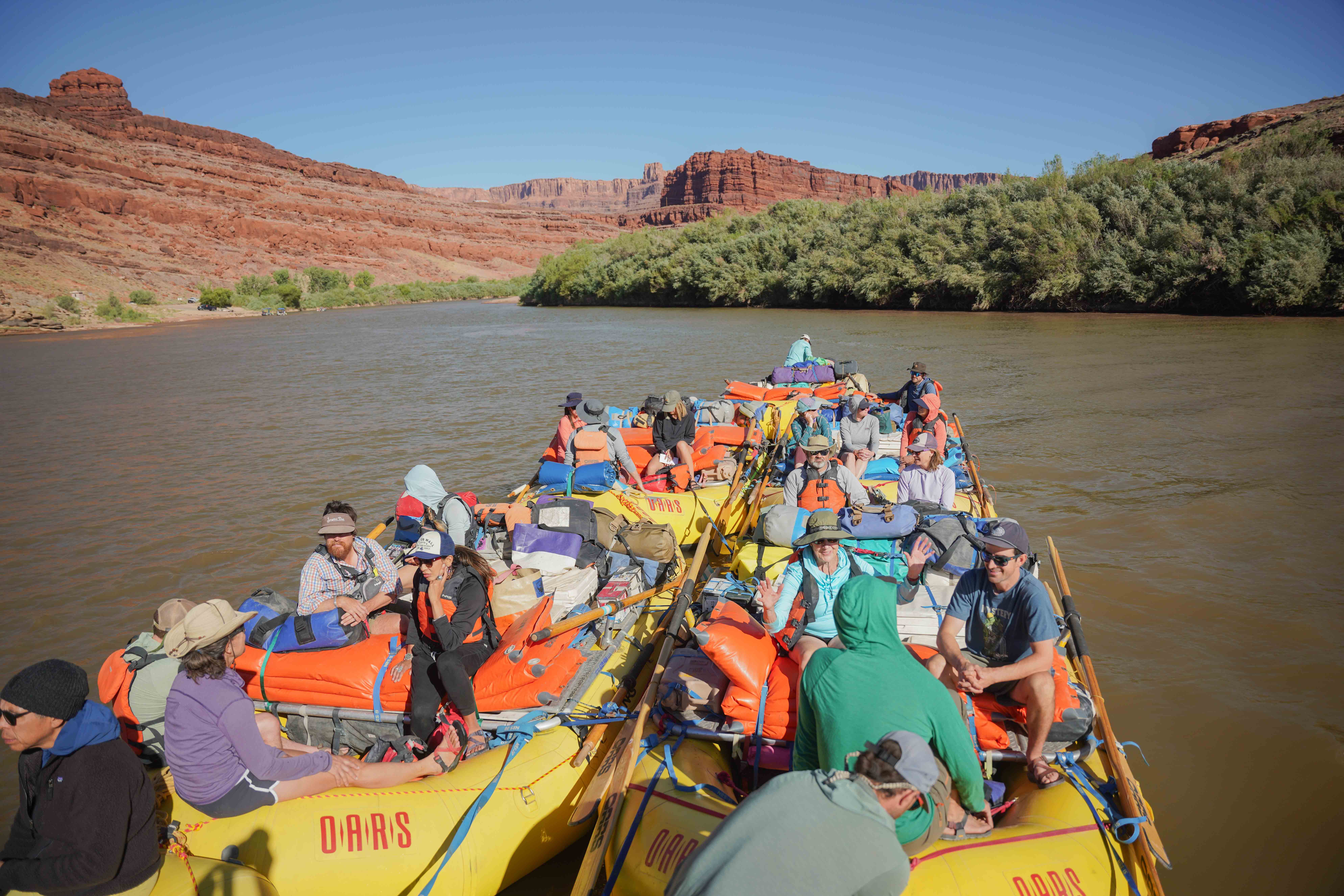 A group of people rafting on a river, surrounded by red rock cliffs and greenery under a clear blue sky.