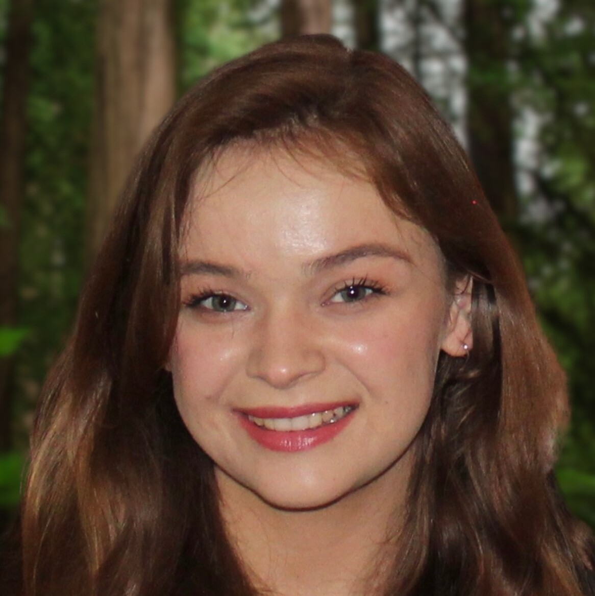 A young woman with long brown hair smiles at the camera, standing in front of tall trees in a forest.