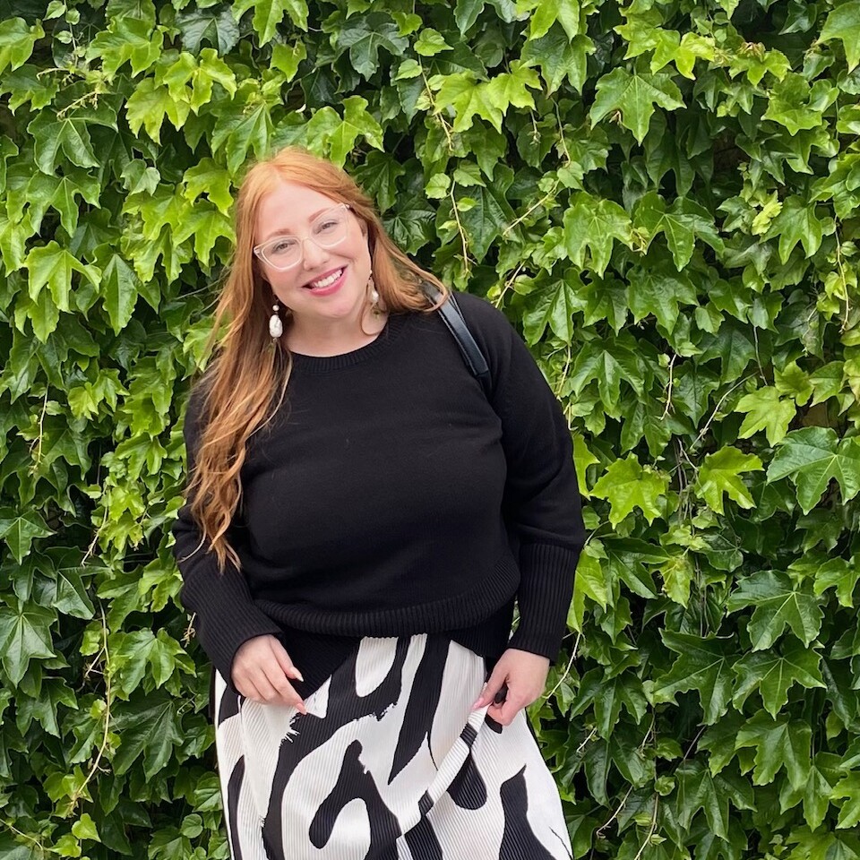 A woman with long red hair wearing glasses, a black sweater, and a patterned skirt stands in front of a leafy green ivy wall, smiling at the camera.