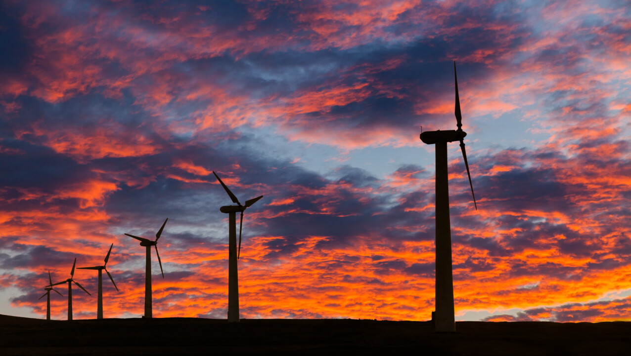 Wind turbines, symbols of climate resilience, are silhouetted against a dramatic sky with vibrant orange and blue clouds at sunset.