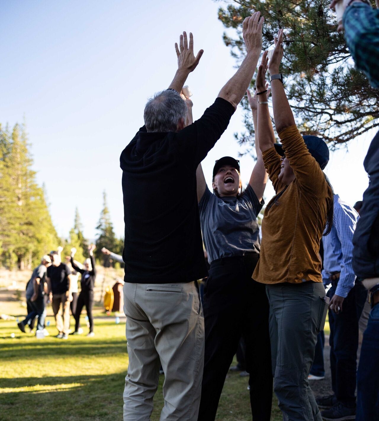 1743297521431 Three people stand on grass outdoors, reaching up and touching hands in a celebratory gesture, with others and trees visible in the background.
