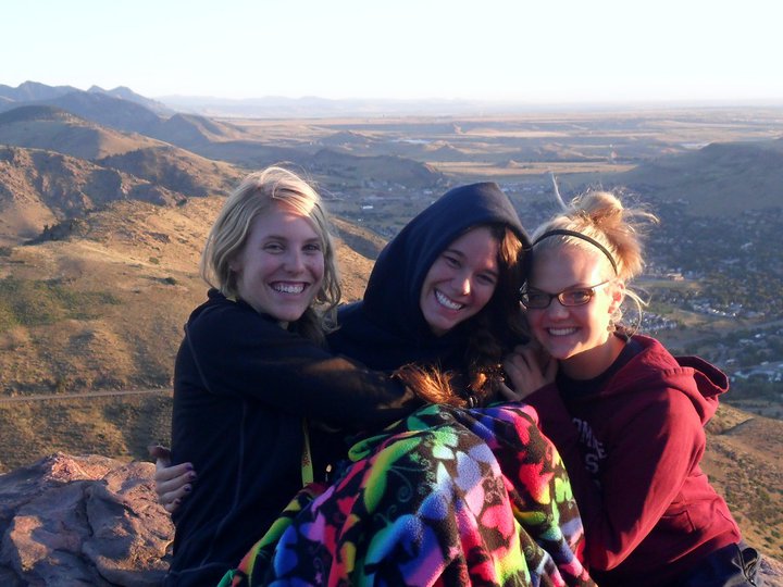 465279043_10161361802223101_3645656827311931792_n Three people sit on a rocky outcrop wrapped in a colorful blanket, smiling with hills and a valley in the background.