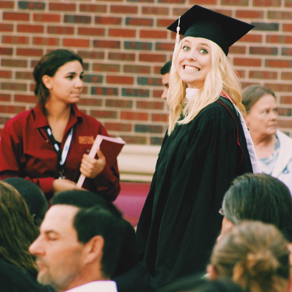 468550433_10161969917752937_5787020048941037350_n A graduate in a cap and gown smiles at the camera during an outdoor graduation ceremony; people are seated and standing around her.