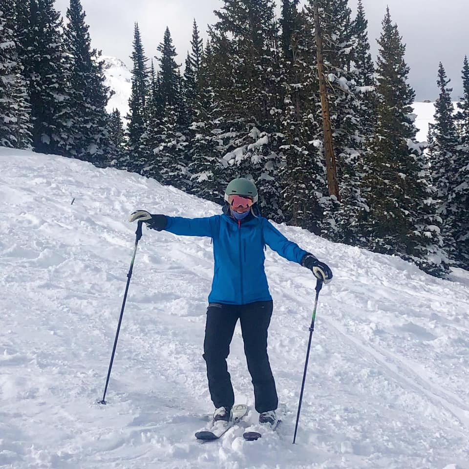 85020176_10157944206782937_4434723659206098944_n A person in winter gear stands on a snowy slope with skis and poles, surrounded by snow-covered pine trees.