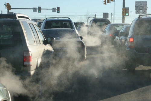 Cars in traffic emit exhaust fumes, creating visible air pollution on a city street under traffic lights.