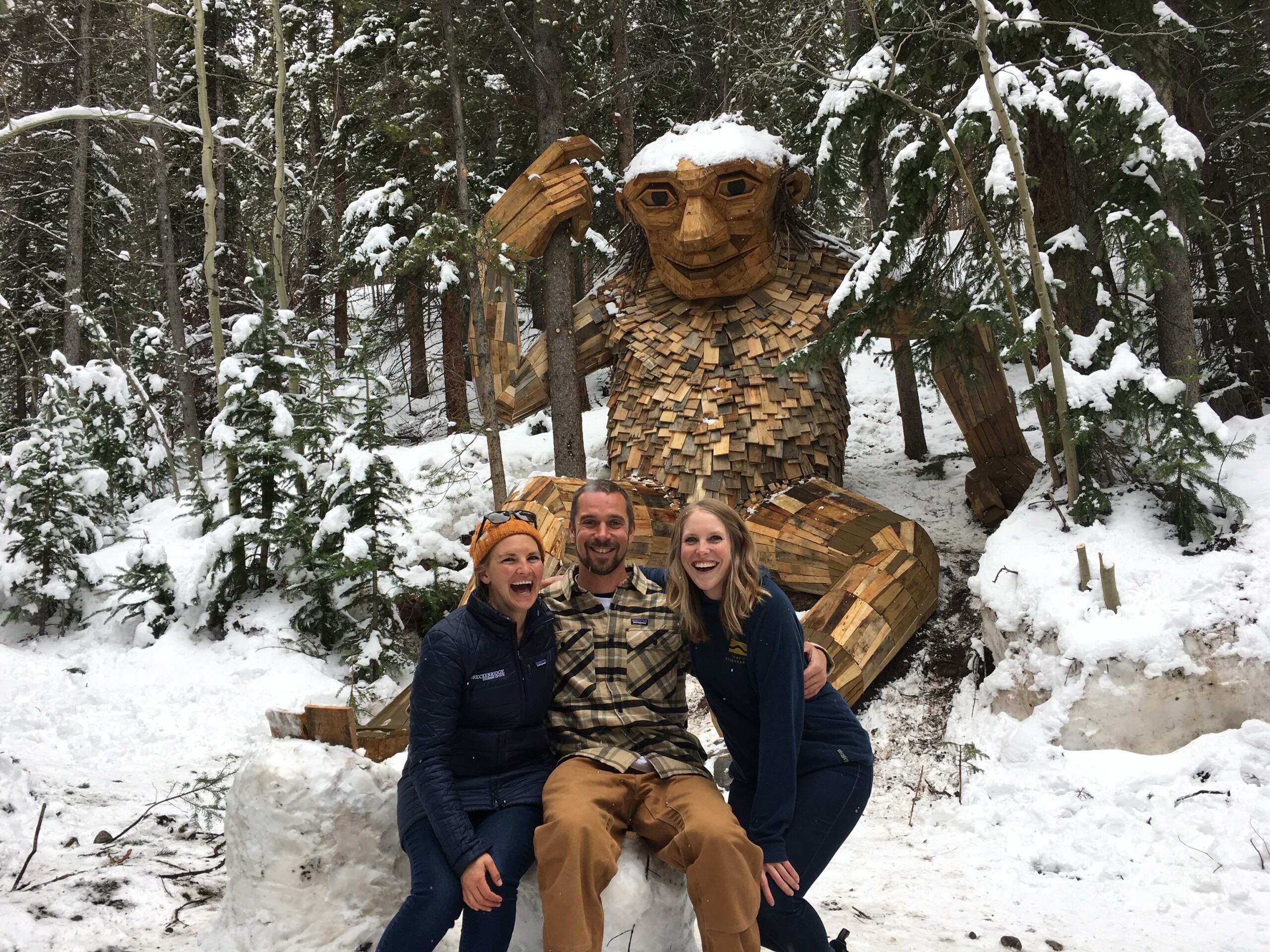 IMG_6558 Three people pose and smile in front of a large wooden troll sculpture in a snowy forest setting.