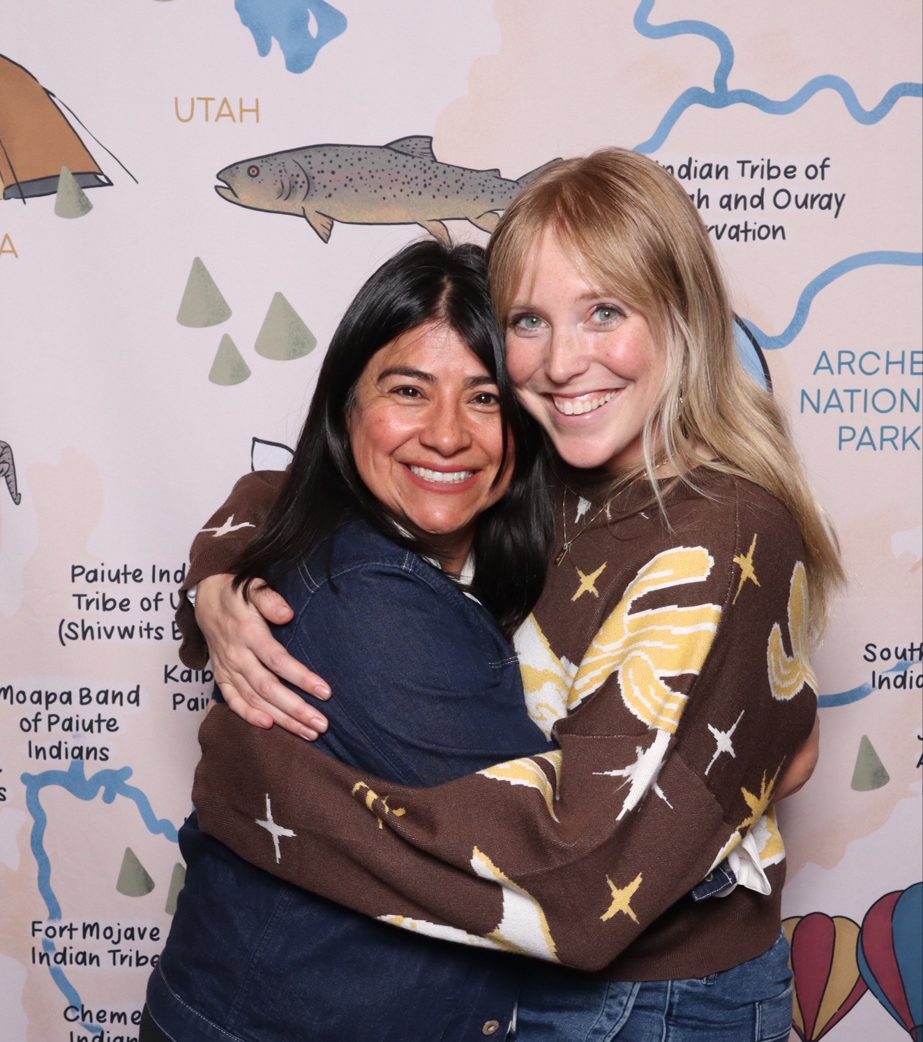 Western_Resource_Advocates_photo_35 Two women smiling and hugging in front of a illustrated map background featuring locations in Utah and surrounding areas.