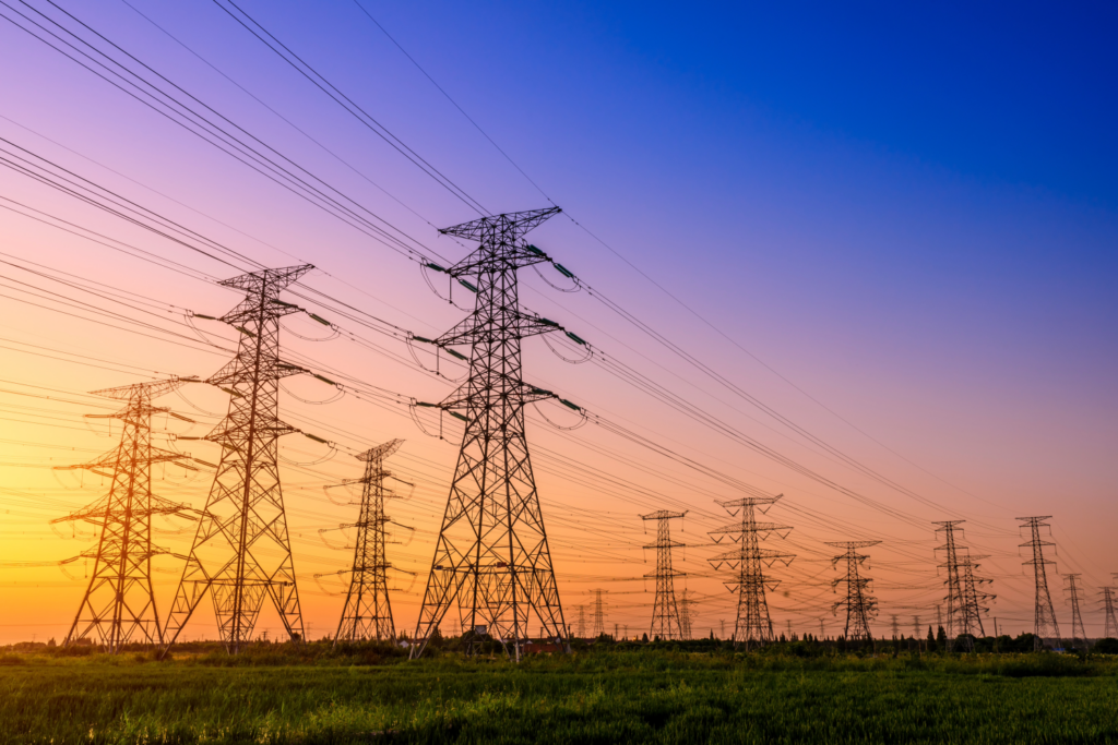Rows of high-voltage power lines and transmission towers stretch across a grassy field under a colorful sunset sky.