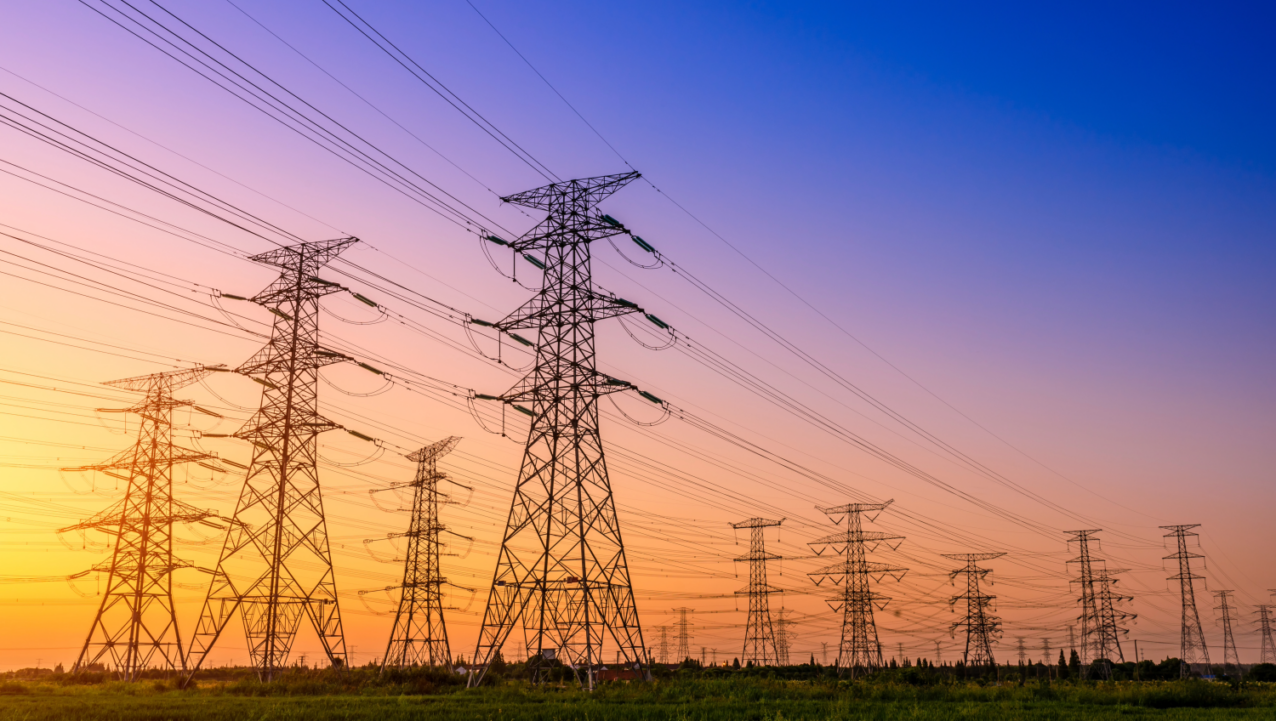 Rows of high-voltage power lines and transmission towers stretch across a grassy field under a colorful sunset sky.
