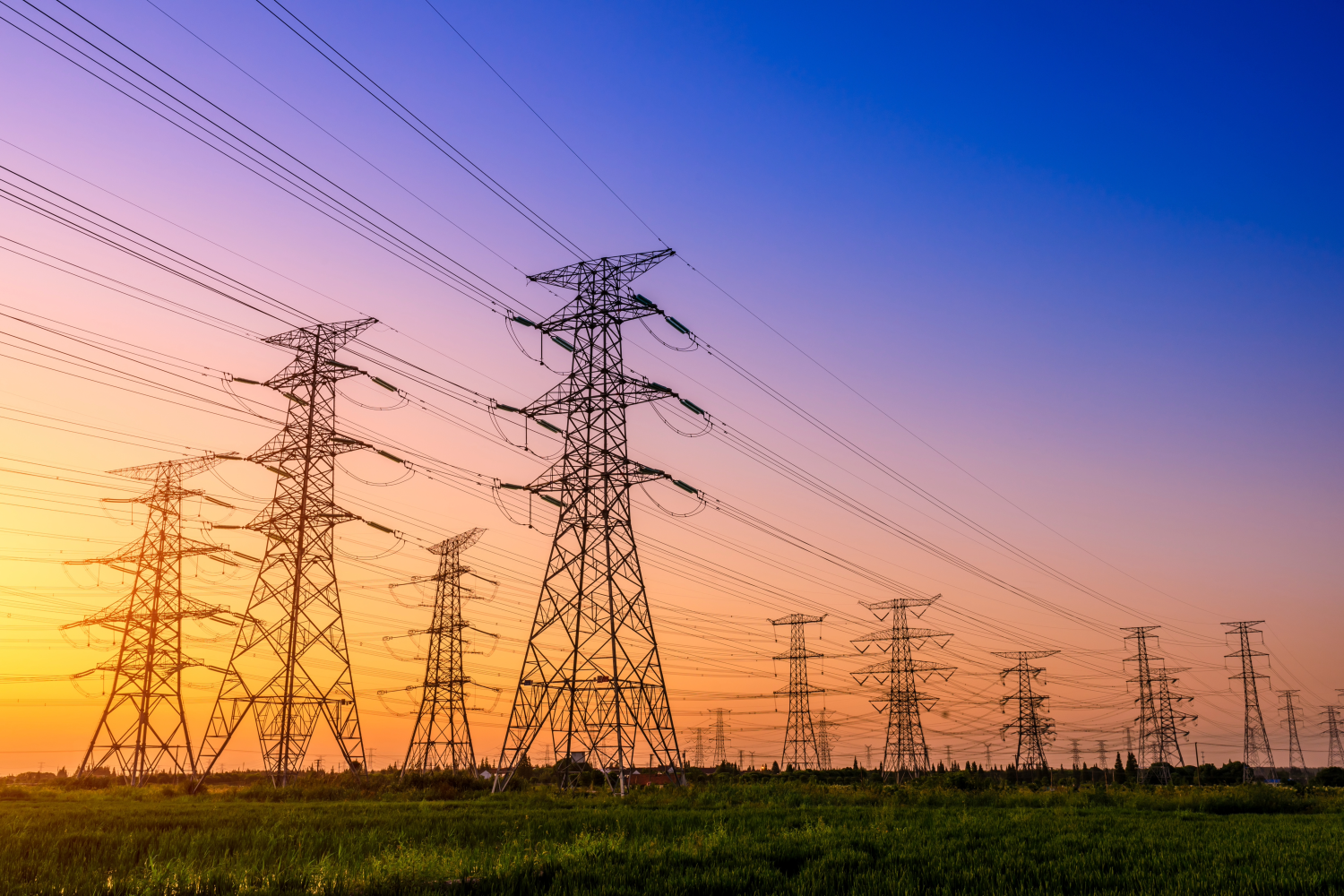 Rows of high-voltage power lines and transmission towers stretch across a grassy field under a colorful sunset sky.