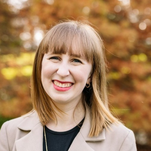 A woman with straight, light brown hair and bangs smiles outdoors, wearing a beige blazer over a black top. Autumn-colored trees are blurred in the background.