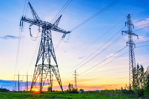 High-voltage power lines and transmission towers stand in a green field at sunset under a partly cloudy sky.