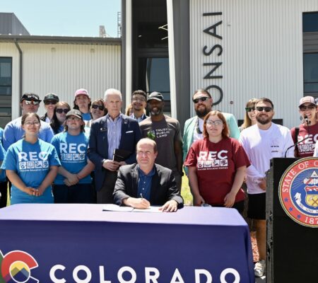 HB25-1215 bill signing - Tarn Udall A group of people pose outdoors around a man signing a document at a table covered with a Colorado banner, next to a podium with the State of Colorado seal.