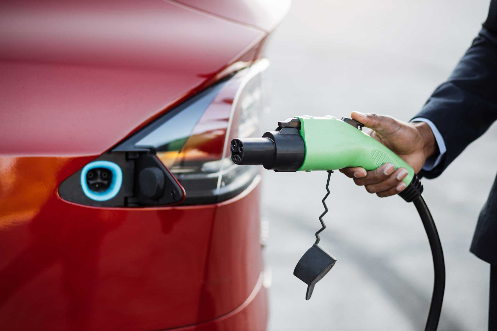 Close up of male hand holding green electric charging plug for luxury red car. African businessman charging his auto on station. Eco friendly transport.